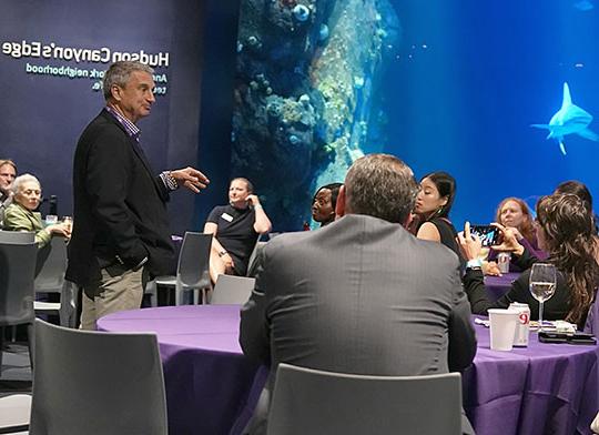 man in sport coat talking to a crowd, standing in front of aquarium